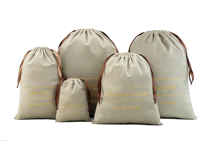 Produktų fotografija studijoje „Subtilūs vizualai“ – penki smėlio spalvos kelioniniai maišeliai su užrašais, skirti drabužiams ir aksesuarams.